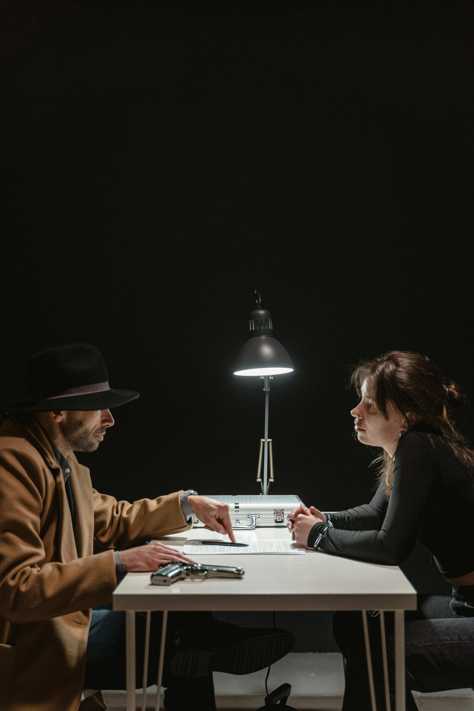 A detective and suspect engaged in a tense interrogation under a single lamp.